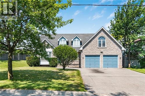Stone facade home featuring a two-car garage with blue doors, a paved driveway, and manicured front lawn with mature trees and shrubs - 381 Kerman Avenue, Grimsby, ON - Outdoor