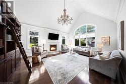 Vaulted ceiling with an ornate chandelier, a large arched window with multiple panes, dark wood-finish flooring, a built-in fireplace with a white mantel, and a tall built-in bookshelf with a rolling - 