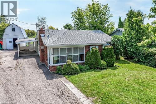 Brick-clad bungalow with a gray shingle roof, featuring a covered carport and a spacious front lawn with mature shrubbery - 114 E Second Road E, Stoney Creek, ON 