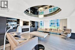 Spacious living area featuring a custom circular ceiling detail with embedded lighting, wood-finish flooring, a wall-mounted fireplace with marble surround, built-in shelving, and large windows - 
