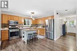 Kitchen featuring wood-finish flooring, light wood cabinetry, stainless steel appliances, a mosaic tile backsplash, and a central island with overhead lighting - 