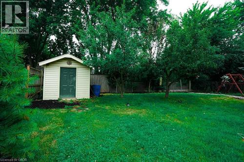 Expansive green lawn featuring a storage shed with beige siding and a dark green door, surrounded by mature trees, a wooden fence, and a swing set structure - 147 Muriel Crescent, London, ON 