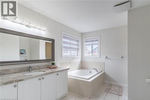 Bathroom featuring a built-in soaking tub with tiled surround, dual windows with blinds, a vanity with a white undermount sink, stone-finish countertop, and a large framed mirror - 47 Hawke Crescent, New Tecumseth, ON 