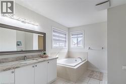 Bathroom featuring a built-in soaking tub with tiled surround, dual windows with blinds, a vanity with a white undermount sink, stone-finish countertop, and a large framed mirror - 