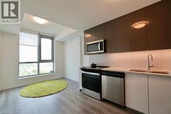 Modern kitchen space featuring wood-finish flooring, sleek cabinetry with under-cabinet lighting, stainless steel appliances, a double basin sink with a gooseneck faucet, and large windows with roller - 