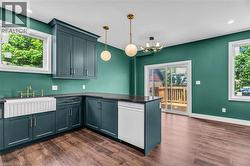 Kitchen featuring deep teal cabinetry, a white fluted farmhouse sink with a brass faucet, dark countertops, and wood-finish flooring - 