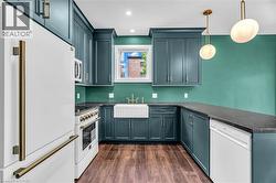 Contemporary kitchen featuring teal cabinetry, dark countertops, wood-finish flooring, a white farmhouse sink, and brass hardware throughout - 