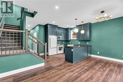 Open-concept living space featuring wood-finish flooring, a contemporary kitchen island with a dark countertop, white appliances, and a staircase with wood newel posts and metal balusters - 