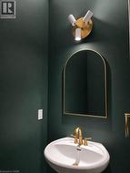 Pedestal sink with brass-finish faucet, arched mirror, and modern wall sconce on a dark green wall - 