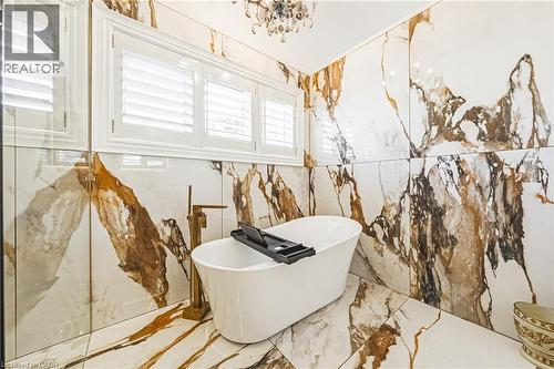Freestanding soaking tub with a floor-mounted brass faucet, surrounded by large format wall and floor tiles featuring white, brown, and gold veining - 712 Highvalley Road, Ancaster, ON 