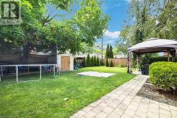 Expansive backyard featuring a large lawn area, mature trees, a paved stone pathway, a wooden storage shed, and a covered gazebo - 