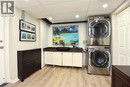 Laundry area featuring a stacked washer and dryer, wood-finish flooring, a utility sink with a dark countertop, white base cabinetry, and recessed lighting - 4299 Lakeshore Road, Burlington, ON - Indoor Photo Showing Laundry Room