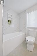 Bathroom featuring a white bathtub with a built-in shower niche, marble-look tile surround, a window with privacy blinds, and light-toned tile flooring - 