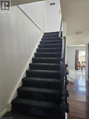Staircase featuring dark carpeted treads and risers, black wooden handrail, and white painted walls - 19 Glenayr Street, Hamilton, ON 
