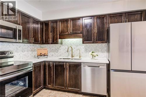Corner kitchen featuring dark wood cabinetry, stainless steel appliances, a white herringbone tile backsplash, and light-colored countertops - 208 Featherstone Crescent, Kitchener, ON - Indoor Photo Showing Kitchen With Double Sink
