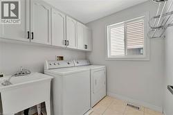 Utility room featuring an overhead bank of white cabinetry with black hardware, a window with horizontal blinds, a utility sink, and tile flooring - 