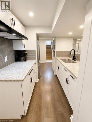 Galley kitchen featuring white cabinetry with black hardware, light-toned countertops, a stainless steel sink with a black faucet, and wood-finish flooring - 355 East 36Th Street, Hamilton, ON 