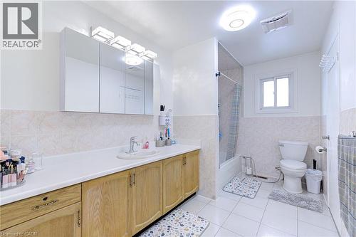 Spacious bathroom featuring a long wood-finish vanity with an integrated sink, a large mirrored medicine cabinet with vanity lighting, a full-size bathtub with an overhead shower, and white tile floor - 214 Merrilee Crescent, Hamilton, ON 