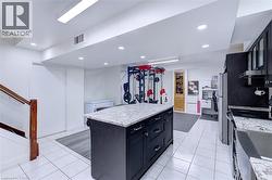 Spacious lower-level featuring a kitchen island with granite countertop and dark cabinetry, white tile flooring, recessed lighting, and a wood-finished staircase with a clear panel railing - 