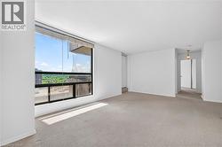 Spacious interior featuring a large window with city and tree-lined scenery, light-toned carpeting, white walls, and an overhead light fixture - 