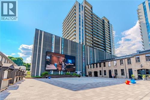 Expansive paver patio with a large outdoor screen, modern high-rise architecture, and adjacent historic brick building - 50 S Grand Avenue S Unit# 1002, Cambridge, ON - Outdoor With Facade