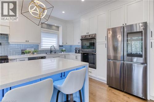 Modern kitchen featuring a large central island with seating, white shaker-style cabinetry, stainless steel appliances, a blue herringbone tile backsplash, and wood-finish flooring - 71 Ian Ormston Drive, Waterloo, ON 