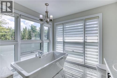 Freestanding soaking tub featuring chrome fixtures, surrounded by large windows with horizontal blinds, polished tile flooring, and a contemporary chandelier - 71 Ian Ormston Drive, Waterloo, ON 