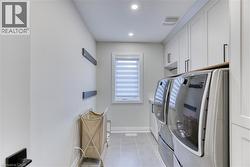 Dedicated laundry space featuring side-by-side front-load units, extensive white cabinetry with black hardware, a light-toned countertop, a window with privacy blinds, and recessed lighting - 