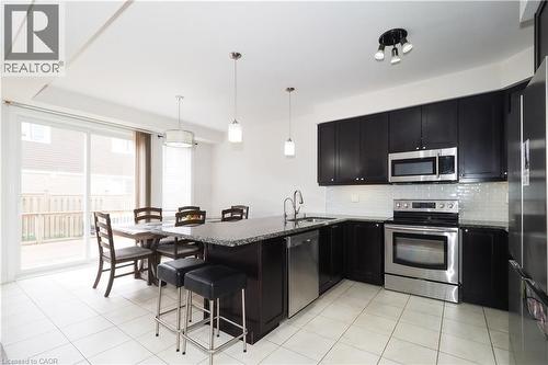 Kitchen featuring dark wood-finish cabinetry, stainless steel appliances, a granite-finish island with seating, a white subway tile backsplash, and ceramic tile flooring - 213 Seabrook Drive, Kitchener, ON 