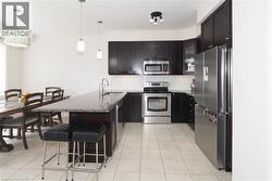 Open-concept kitchen featuring dark wood cabinetry, stainless steel appliances, a center island with a stone-finish countertop, and tile flooring - 