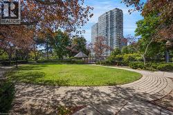 Expansive green space featuring a circular paver walkway, mature trees, manicured shrubs, a wooden gazebo, and a multi-story building in the background - 