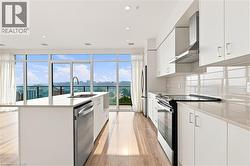 Open-concept kitchen with expansive floor-to-ceiling windows, a large center island featuring an undermount sink and stainless steel dishwasher, wood-finish flooring, and a full suite of stainless ste - 