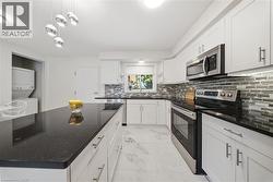 Modern kitchen featuring an expansive center island with dark countertops, white cabinetry, stainless steel appliances, a mosaic tile backsplash, and light-toned tile flooring - 