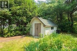 Storage shed with vinyl siding, a gambrel roof, and a single window set amidst mature trees and lush landscaping - 