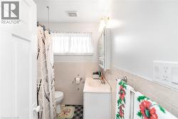 Full bathroom featuring a window with privacy curtain, white vanity with integrated sink, wall-mounted medicine cabinet with brass hardware, and a ceramic tile wainscoting - 