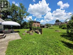 Expansive grassy yard with mature trees, featuring a stone patio, a small storage shed, and a designated fire pit area with tree stump seating - 