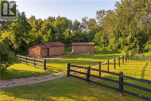 Expansive grassy area featuring a dark-stained split-rail fence, two wooden outbuildings with reddish-brown roofs, and a backdrop of mature trees - 12613 Second Line, Milton, ON - Outdoor