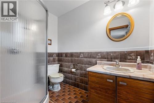 Bathroom featuring a shower stall with fluted glass doors, a vanity with wood-finish cabinetry, a marble-patterned countertop, a decorative oval mirror, and ceramic floor tiling - 7 Thomson Road, Simcoe, ON - Indoor Photo Showing Bathroom