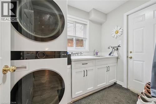 Laundry area featuring a stacked washer and dryer, white cabinetry with black hardware, a stainless steel sink, a window with blinds, and light-colored walls - 916 Rush Meadow Court, Kitchener, ON - Indoor Photo Showing Other Room