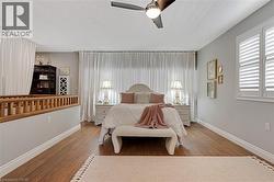 Bedroom featuring wood-finish flooring, plantation shutters, full-height white drapery, a ceiling fan with integrated lighting, and a partial wood railing - 