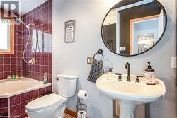 Bathroom featuring a pedestal sink with a dark-finish faucet, a circular mirror, and a built-in corner bathtub with maroon tile surround - 