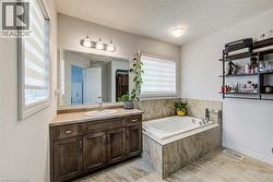 Bathroom featuring a built-in soaking tub with stone-look tile surround, dual windows with privacy blinds, and a wood-finish vanity with an integrated sink and countertop - 