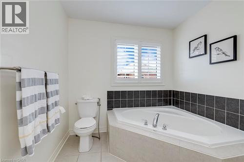 Corner soaking tub with polished chrome fixtures, white window shutters, dark tile wainscoting, white toilet, and neutral floor tiling - 598 Bessborough Drive, Milton, ON - Indoor Photo Showing Bathroom
