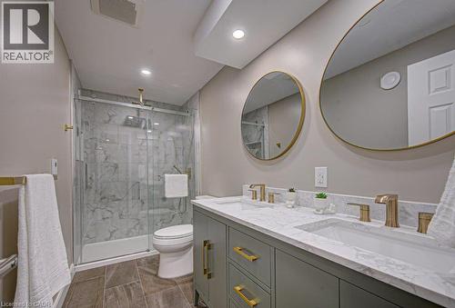 Dual vanity with brass hardware and round mirrors, a glass-enclosed shower with a rainfall showerhead, full-height marble-patterned tile, and wood-finish flooring - 15 Matheson Crescent, Innerkip, ON - Indoor Photo Showing Bathroom