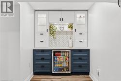 Built-in beverage station featuring white upper cabinetry with glass-front accents, contrasting blue lower cabinetry, a white countertop, and a tiled backsplash - 