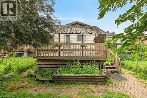 Rear exterior featuring a wooden deck with railing and stairs, light-colored siding, and a raised garden bed with a wooden border - 23 Harrison Avenue, Hamilton, ON - Outdoor With Deck Patio Veranda
