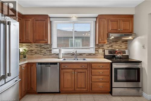 Kitchen featuring warm wood cabinetry, stainless steel appliances, a mosaic tile backsplash, a double basin sink, and neutral tile flooring - 56 Brookstream Court, Hamilton, ON - Indoor Photo Showing Kitchen With Stainless Steel Kitchen With Double Sink