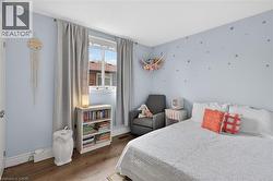 Bedroom featuring light blue walls, wood-finish flooring, a double window with light-filtering curtains, white baseboards, and a built-in wall heater - 