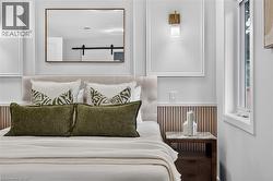 Bedroom featuring wainscoting, an upholstered headboard, a wall-mounted brass light fixture, and a framed mirror - 