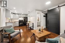 Open-concept living area featuring wood-finish flooring, a contemporary kitchen with light grey cabinetry, black appliances, and a butcher block style countertop - 